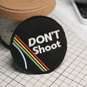 May include: Black, round embroidered patch with the words "DON'T Shoot" in white. A rainbow design of blue, yellow, and red lines is on the left side. The patch is on a grid surface.