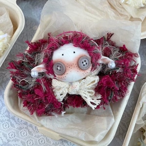 May include: A handcrafted doll with a white face, button eyes, and a crocheted bow. The doll features red and grey yarn hair and is displayed in a small, white container. The doll's cheeks are blushed with pink.