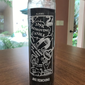 May include: A black candle with white text and images. The text reads "Alleged Jinx Removing Candle". The images include a snake, a horseshoe, a heart, dice, a cat, and a hand. The candle is on a wooden surface.