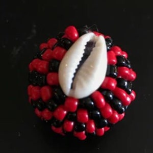 May include: A small, round bead made of black and red beads with a white cowrie shell in the center.