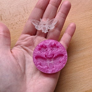 Puede incluir: Un molde de silicona rosa con un diseño en relieve de dos polillas. El molde está en la palma de una mano.