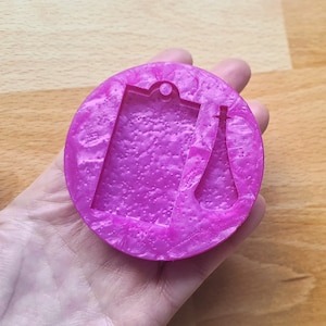 Könnte beinhalten: Eine rosa Harzform mit zwei Formen: ein Etikett mit einem Loch oben und eine Flasche mit abgerundetem Oberteil. Die Form hat eine gesprenkelte, marmorierte Optik.