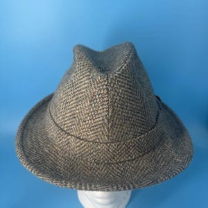 May include: A brown and grey fedora hat featuring a classic herringbone pattern throughout the fabric. The hat has a structured crown with a central crease and a curved brim that encircles the base, showcasing a textured wool-like weave.