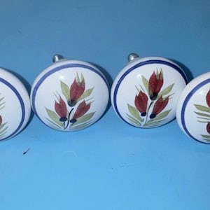 May include: Four white ceramic cabinet knobs with a floral design. Each knob features a red and green floral pattern on a white background, framed by a blue and white border. The knobs are set against a blue backdrop.