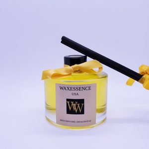 May include: A clear glass reed diffuser bottle filled with yellow liquid, labelled "Waxessence USA." The bottle has a black cap, a yellow ribbon, and black reeds. The label also says "Reed Diffuser | 200 ml (6.8 fl oz)."