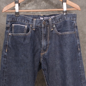 Puede incluir: Vaqueros de mezclilla azul oscuro colgados de una percha de madera. Los vaqueros tienen un diseño clásico de cinco bolsillos, cierre de botón y cremallera, y la marca "GAP STANDARD TAPER" es visible en la cintura. Las costuras son de color naranja en contraste.