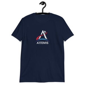 Puede incluir: Una camiseta azul marino con el logotipo del programa Artemis. El logotipo presenta una letra A estilizada blanca con una órbita roja y azul a su alrededor. El texto "ARTEMIS" está debajo del logotipo.