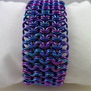 May include: A wide bracelet made of interlocking metal rings. The bracelet features a color-shifting finish, with hues of blue, purple, and teal. The chainmail-style design is displayed on a white surface.