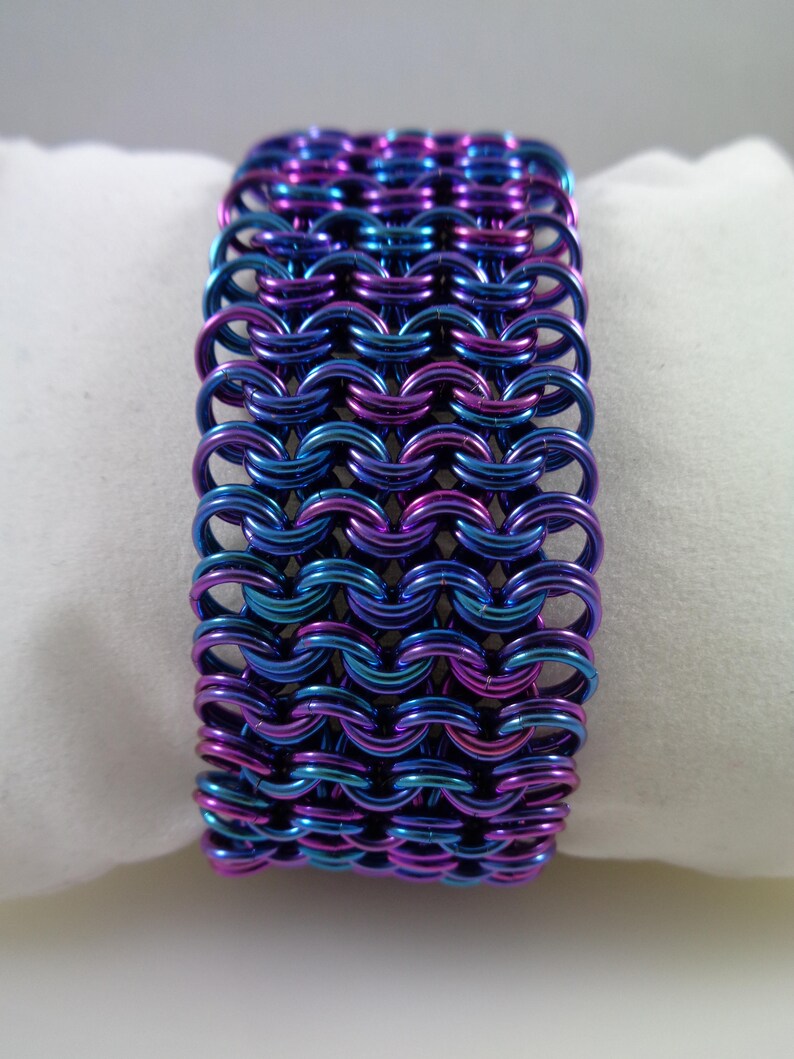 May include: A wide bracelet made of interlocking metal rings. The bracelet features a color-shifting finish, with hues of blue, purple, and teal. The chainmail-style design is displayed on a white surface.