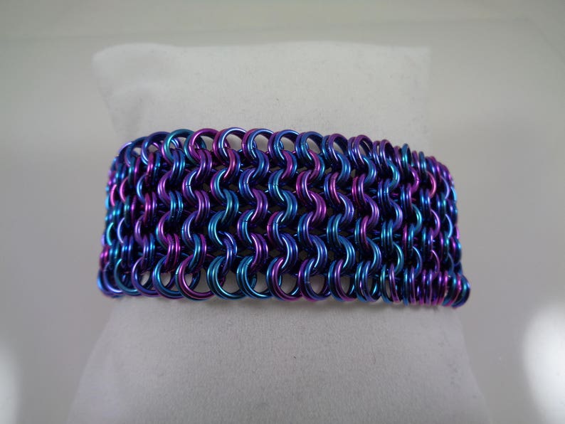 May include: A wide bracelet made of interlocking metal rings. The bracelet features a color-shifting effect, with hues of blue, purple, and pink. The chainmail-style design is displayed on a white surface.