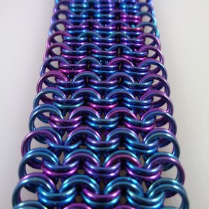 May include: Close-up of a section of chainmail in shades of blue and purple. The metal rings are interlocked in a dense pattern, creating a textured surface. The colors shift with the light, giving the chainmail a shimmering appearance.