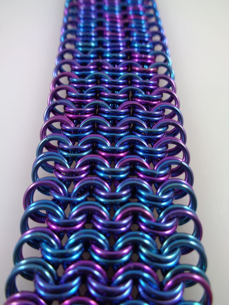 May include: Close-up of a section of chainmail in shades of blue and purple. The metal rings are interlocked in a dense pattern, creating a textured surface. The colors shift with the light, giving the chainmail a shimmering appearance.