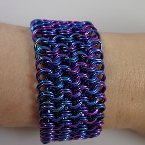 May include: A wide bracelet made of interlocking metal rings. The bracelet is a gradient of blue and purple hues. The jewelry is worn on a wrist, showcasing the intricate chainmail design. The bracelet is approximately 2 inches wide.