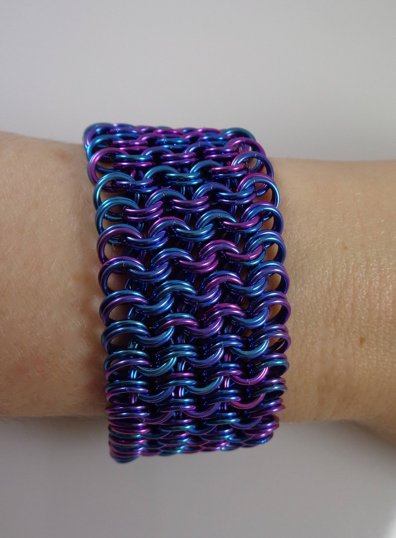 May include: A wide bracelet made of interlocking metal rings. The bracelet is a gradient of blue and purple hues. The jewelry is worn on a wrist, showcasing the intricate chainmail design. The bracelet is approximately 2 inches wide.