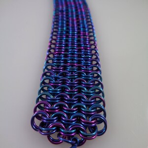 May include: A chainmail bracelet in shades of blue, purple, and teal. The metal links are intricately woven together, creating a flexible band. A matching clasp secures the bracelet.