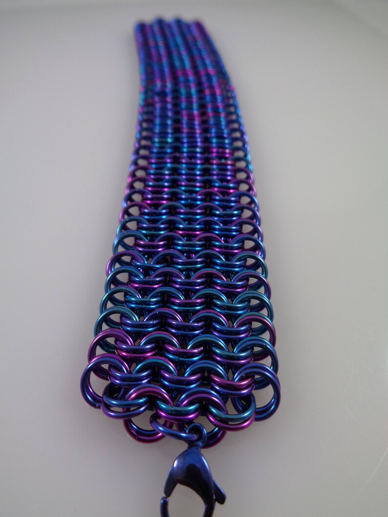 May include: A chainmail bracelet in shades of blue, purple, and teal. The metal links are intricately woven together, creating a flexible band. A matching clasp secures the bracelet.