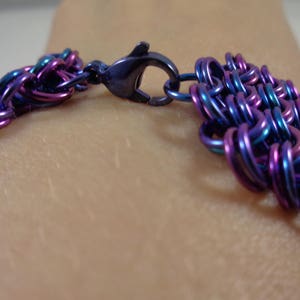May include: Close-up of a bracelet with a woven design. The bracelet is made of small, iridescent metal rings in shades of blue, purple, and teal. A dark blue clasp secures the bracelet.