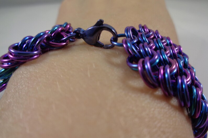 May include: Close-up of a bracelet with a woven design. The bracelet is made of small, iridescent metal rings in shades of blue, purple, and teal. A dark blue clasp secures the bracelet.