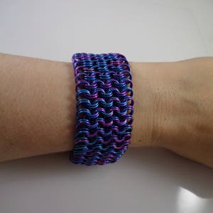 May include: A wide bracelet made of interlocking metal rings, showcasing a vibrant color gradient of blue, purple, and teal. The chainmail-style bracelet is worn on a wrist, offering a bold and eye-catching accessory.