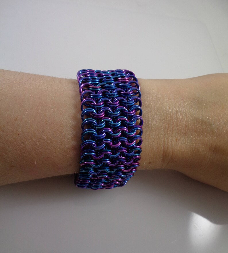 May include: A wide bracelet made of interlocking metal rings, showcasing a vibrant color gradient of blue, purple, and teal. The chainmail-style bracelet is worn on a wrist, offering a bold and eye-catching accessory.