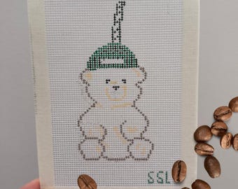 May include: A hand-stitched needlepoint featuring a teddy bear wearing a green hat with a black and white design. The bear is outlined in a light brown color. The letters "SSL" are in green. Coffee beans are scattered around the needlepoint.