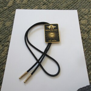 May include: Black bolo tie with a gold-colored metal charm featuring the text "Shrine Odyssey 2002 Las Vegas".