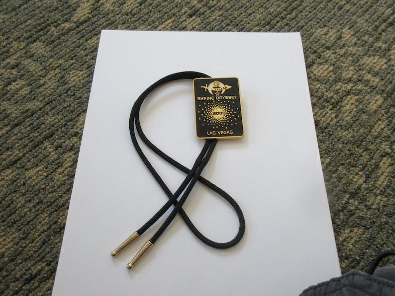 May include: Black bolo tie with a gold-colored metal charm featuring the text "Shrine Odyssey 2002 Las Vegas".