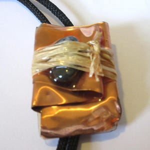 May include: A copper bolo tie with a layered design and a dark blue stone wrapped in tan raffia.