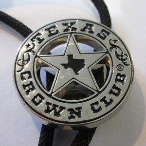 Puede incluir: Corbata bolo de plata con una estrella negra y plateada que presenta el contorno de Texas. La corbata tiene las palabras "Texas Crown Club" alrededor de la estrella.