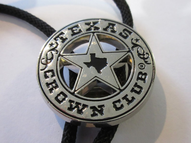 Puede incluir: Corbata bolo de plata con una estrella negra y plateada que presenta el contorno de Texas. La corbata tiene las palabras "Texas Crown Club" alrededor de la estrella.