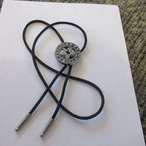 Puede incluir: Un corbat&iacute;n bolo negro con un colgante de metal plateado que presenta una estrella de cinco puntas y el texto "Texas Crown Club".