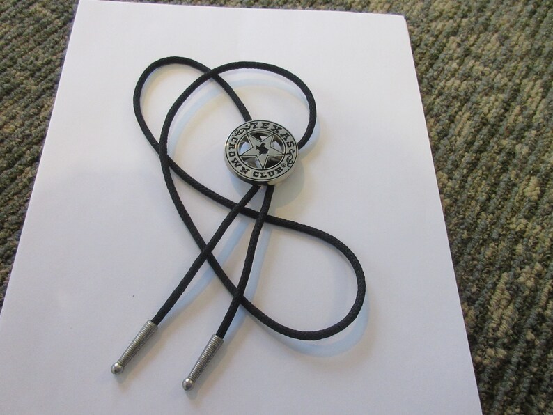 Puede incluir: Un corbat&iacute;n bolo negro con un colgante de metal plateado que presenta una estrella de cinco puntas y el texto "Texas Crown Club".