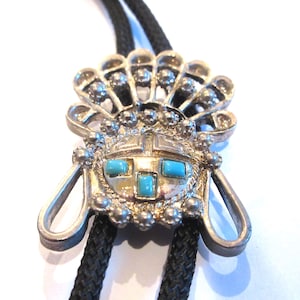 May include: Silver-toned bolo tie with a Native American-inspired design. The pendant features a headdress motif with turquoise-colored stone accents and a black cord. The bolo tie is a unique accessory.