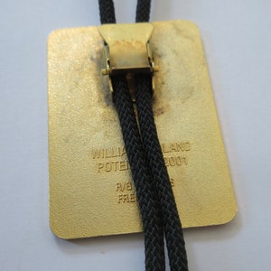 May include: A gold-colored bolo tie with a black cord and a rectangular metal slide. The slide has the text "WILLIAM POTTERLAND 2001" engraved on it.