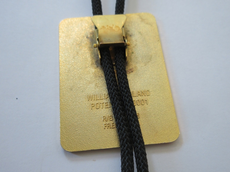 May include: A gold-colored bolo tie with a black cord and a rectangular metal slide. The slide has the text "WILLIAM POTTERLAND 2001" engraved on it.