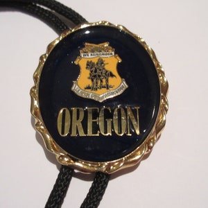 May include: A gold bolo tie with a blue and gold circular pendant featuring the state seal of Oregon. The seal depicts a rider on horseback with the words "We Remember" and "The State of Oregon" on a yellow shield. The word "OREGON" is written in gold letters on the pendant.