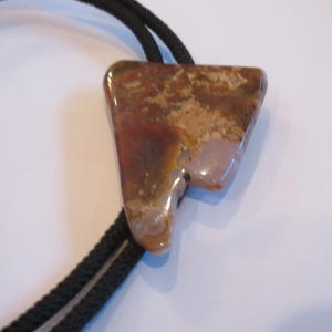 May include: A bolo tie featuring a polished, triangular stone pendant with a mix of brown, tan, and cream colors. The stone is attached to a black cord with silver-colored tips, creating a classic Western accessory.