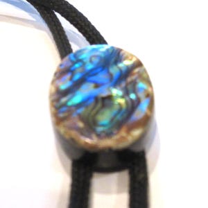 May include: Oval-shaped bolo tie slide with iridescent abalone shell inlay, showcasing blues, greens, and golds. The slide is set on a black braided cord, creating a striking contrast. A unique accessory.