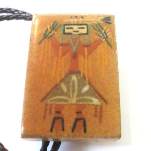 May include: A rectangular bolo tie slide featuring a tan background with a painted figure in orange, black, and white. The figure has a square head, raised arms holding stalks, and a triangular skirt. A dark brown braided leather cord is attached.