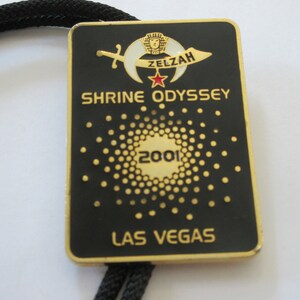 May include: Black and gold metal bolo tie with a black and white crescent moon and star design. The tie features the text "Zelzah Shrine Odyssey 2001 Las Vegas".