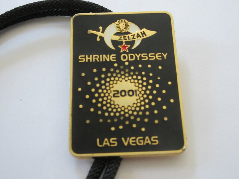 May include: Black and gold metal bolo tie with a black and white crescent moon and star design. The tie features the text "Zelzah Shrine Odyssey 2001 Las Vegas".