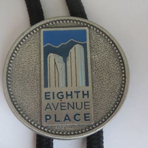 May include: A silver bolo tie with a round, textured metal pendant. The pendant features a blue and white illustration of two buildings with the text "EIGHTH AVENUE PLACE".
