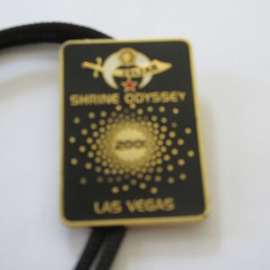 May include: A black and gold metal bolo tie with a black background and gold lettering. The text reads "Shrine Odyssey 200X Las Vegas". The bolo tie features a gold crescent moon and star with a red jewel in the center.
