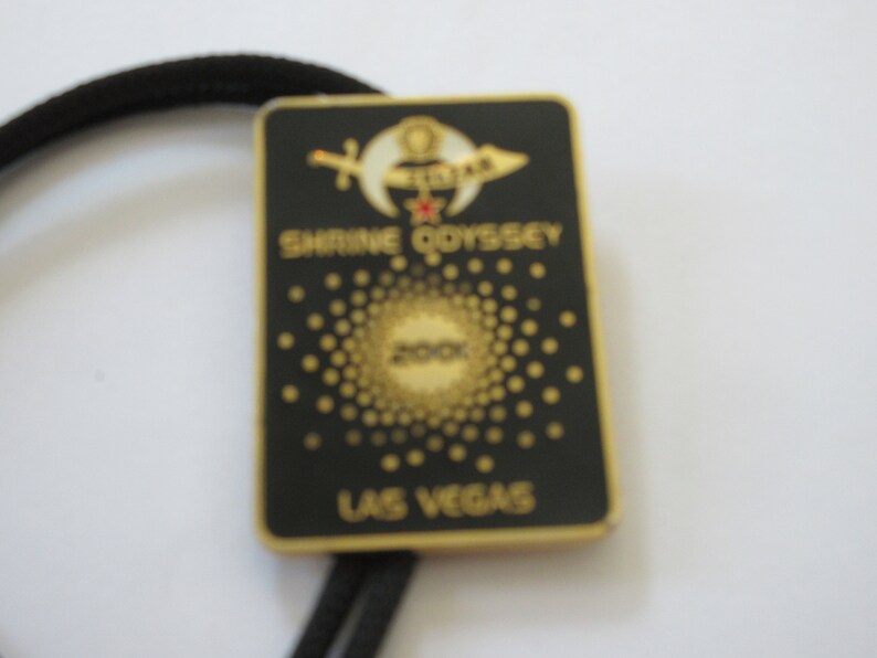 May include: A black and gold metal bolo tie with a black background and gold lettering. The text reads "Shrine Odyssey 200X Las Vegas". The bolo tie features a gold crescent moon and star with a red jewel in the center.
