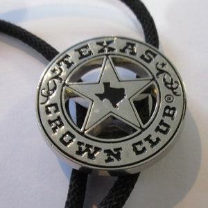 Puede incluir: Un bolo tie plateado con un dije en forma de estrella negro y plateado. El dije presenta el contorno del estado de Texas dentro de la estrella y las palabras "Texas Crown Club" alrededor de la estrella.