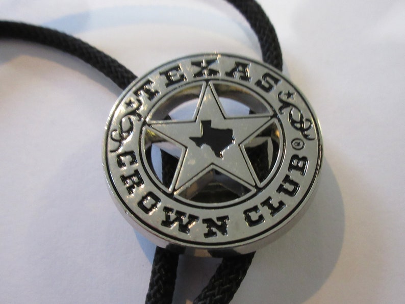 Puede incluir: Un bolo tie plateado con un dije en forma de estrella negro y plateado. El dije presenta el contorno del estado de Texas dentro de la estrella y las palabras "Texas Crown Club" alrededor de la estrella.