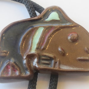 May include: A brown ceramic bolo tie featuring a stylized design of a fish with a green and red accent. The tie is attached to a black cord.