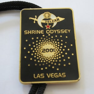 May include: A black and gold metal bolo tie with a gold-colored emblem featuring a crescent moon, a star, and a sword. The text "ZELZAH SHRINE ODYSSEY 2001 LAS VEGAS" is printed on the emblem.