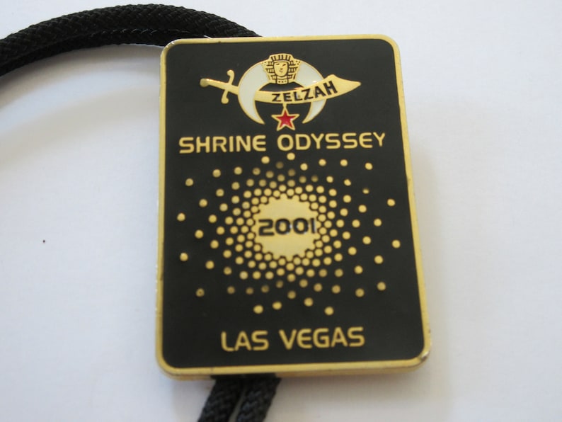 May include: A black and gold metal bolo tie with a gold-colored emblem featuring a crescent moon, a star, and a sword. The text "ZELZAH SHRINE ODYSSEY 2001 LAS VEGAS" is printed on the emblem.