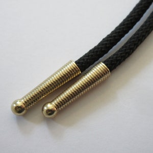 May include: Close-up of a black bolo tie with gold-toned metal tips. The tips have a ridged design.